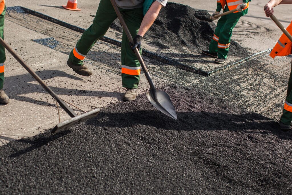 Workers in safety gear spread and level fresh asphalt on a road with shovels and rakes during a construction or repair project, providing top-notch asphalt services New Hampshire can rely on.