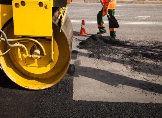 A worker spreads asphalt with a shovel while a steamroller compacts the new pavement on a road under construction.