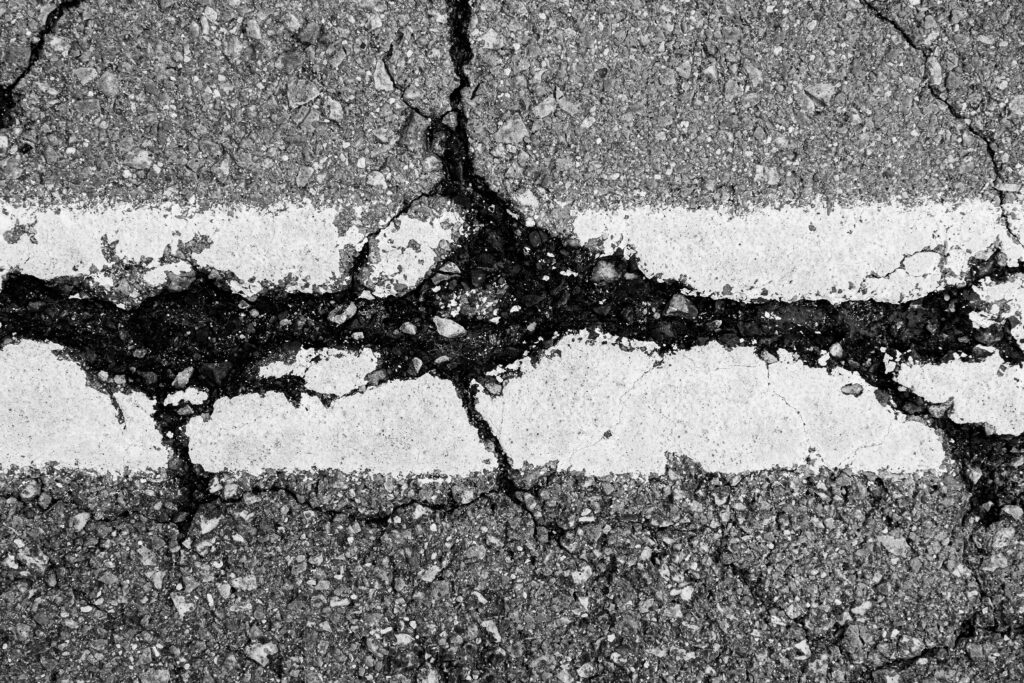 Close-up of a cracked asphalt road with two parallel white lines, highlighting significant damage and rough texture—ideal for crack sealing New Hampshire roads. Close-up of a cracked asphalt road with two parallel white lines, highlighting significant damage and rough texture—ideal for crack sealing New Hampshire roads.