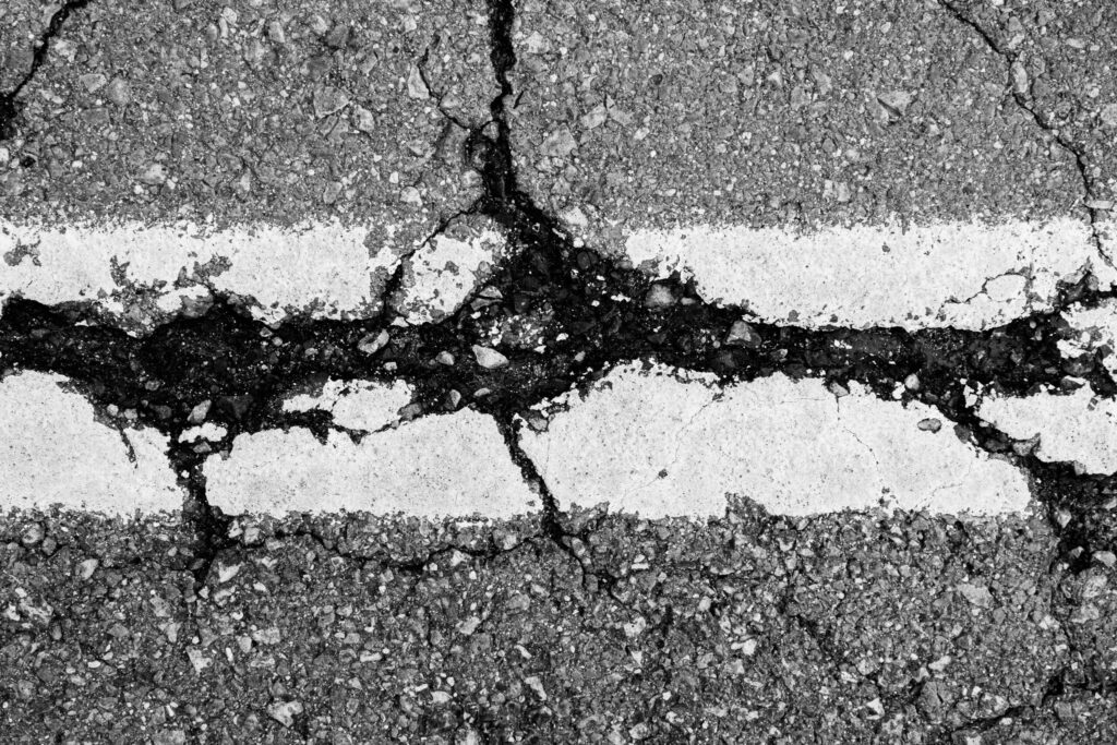 Close-up of a cracked asphalt road with two parallel white lines, highlighting significant damage and rough texture—ideal for crack sealing New Hampshire roads.