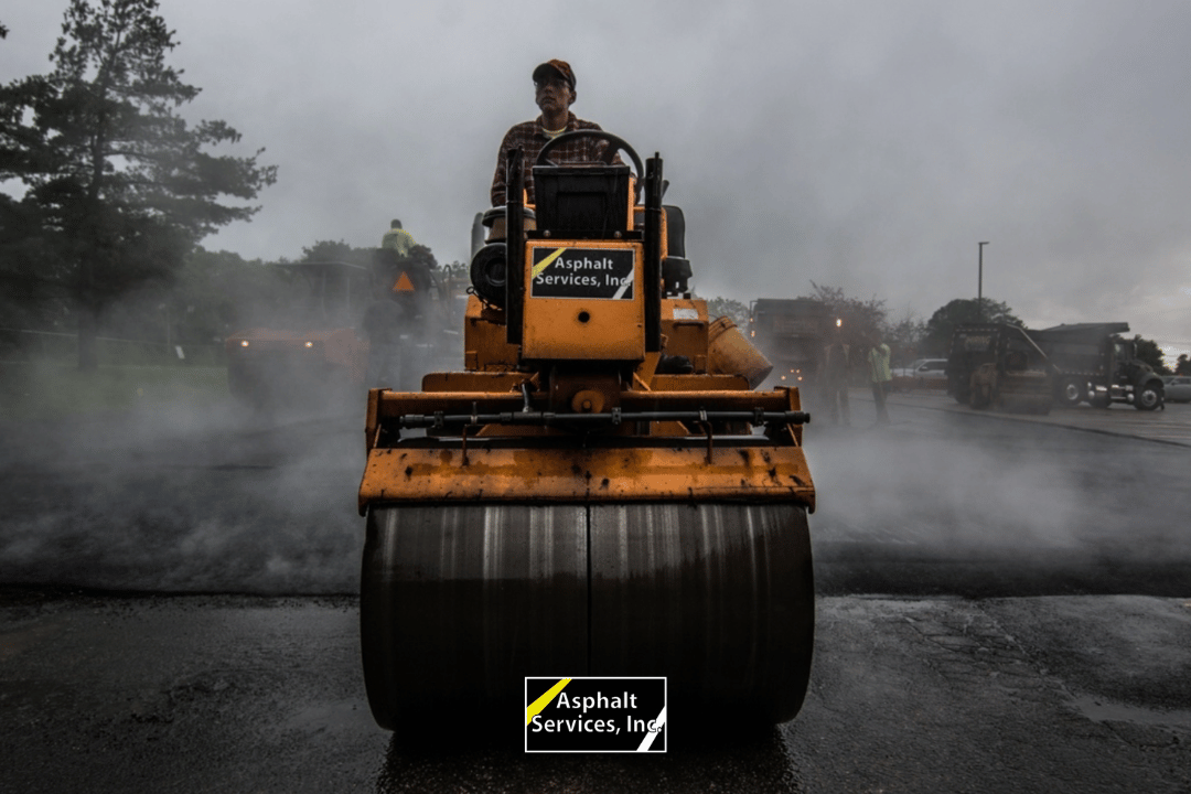 A worker operates a yellow asphalt roller on a steaming road, with "Asphalt Services, Inc." displayed on the machine; other workers and machinery are seen in the misty background.