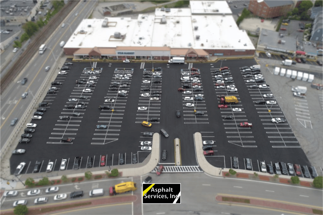 Aerial view of a large, freshly paved parking lot with parked cars in front of a supermarket; text reads "Asphalt Services, Inc." near the entrance.