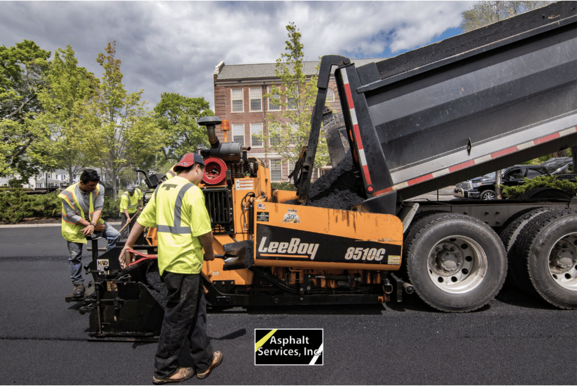 Workers of top paving contractor Asphalt Services Inc. operate paving equipment next to a dump truck pouring asphalt onto a road in front of a brick building, with trees and greenery in the background.