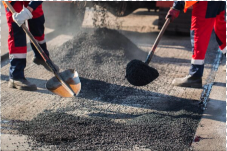 Workers in safety gear use shovels to spread fresh asphalt on a road surface during daylight, ensuring even coverage, a process often estimated beforehand with an Asphalt Driveway Cost Calculator.