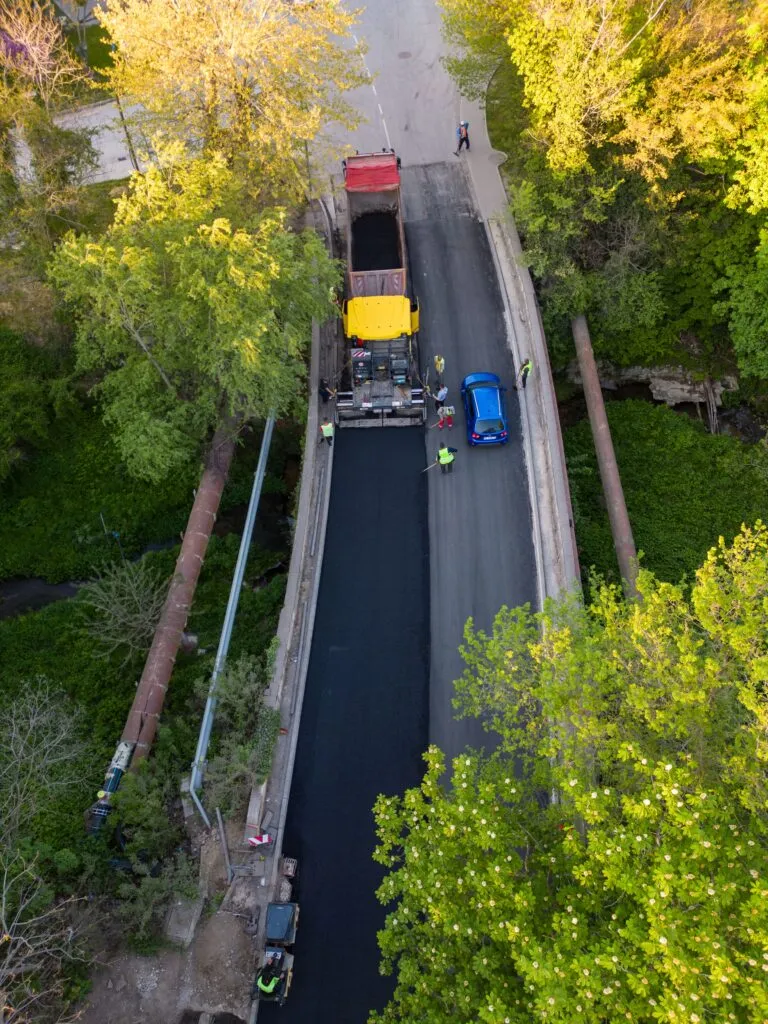 Aerial view of a road construction crew paving a bridge with asphalt. Visible are construction vehicles, workers in safety vests, and a blue car waiting to cross. Surrounded by green trees.