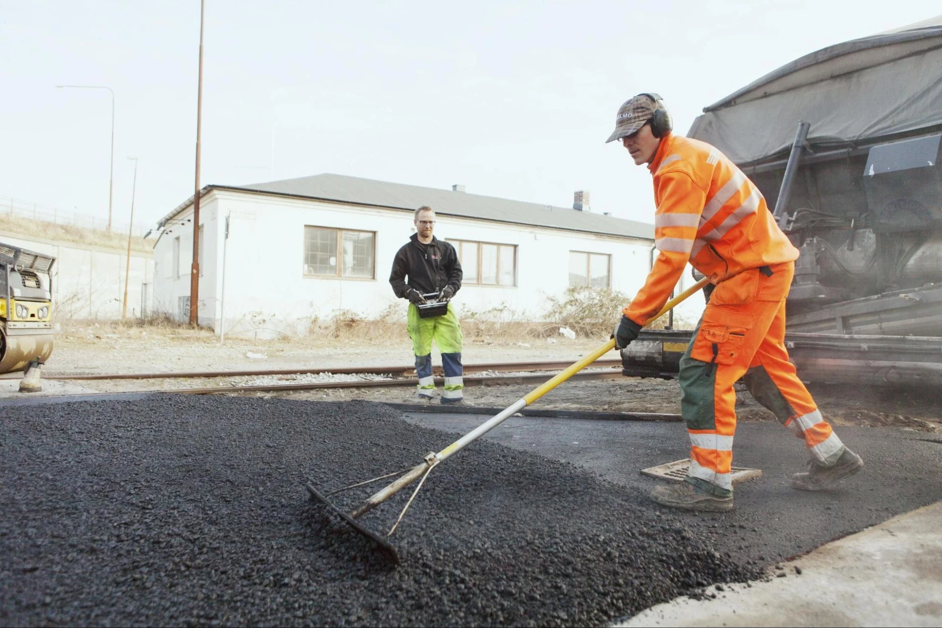 Workers in high-visibility clothing diligently provide asphalt services on a road near a building. One expertly uses a rake while the other oversees the process. A vehicle is clearly visible in the background, completing the scene of efficient teamwork. Workers in high-visibility clothing diligently provide asphalt services on a road near a building. One expertly uses a rake while the other oversees the process. A vehicle is clearly visible in the background, completing the scene of efficient teamwork.