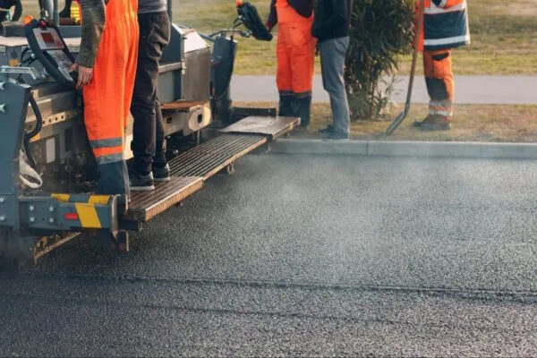 Workers in orange uniforms operate paving machinery on a road. Steam rises from freshly laid asphalt.