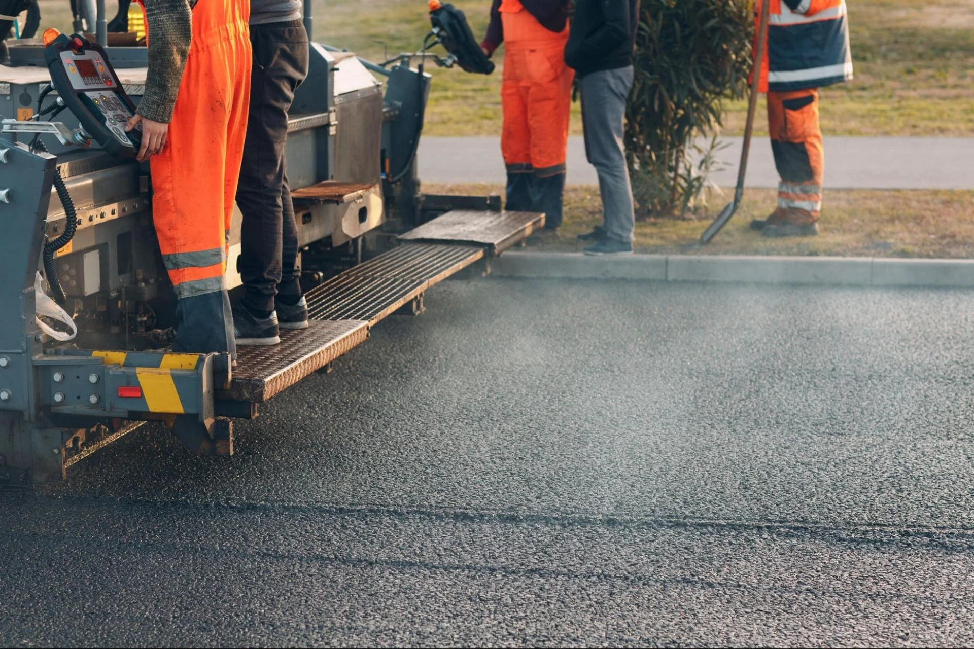 Workers in orange uniforms operate paving machinery on a road. Steam rises from freshly laid asphalt.