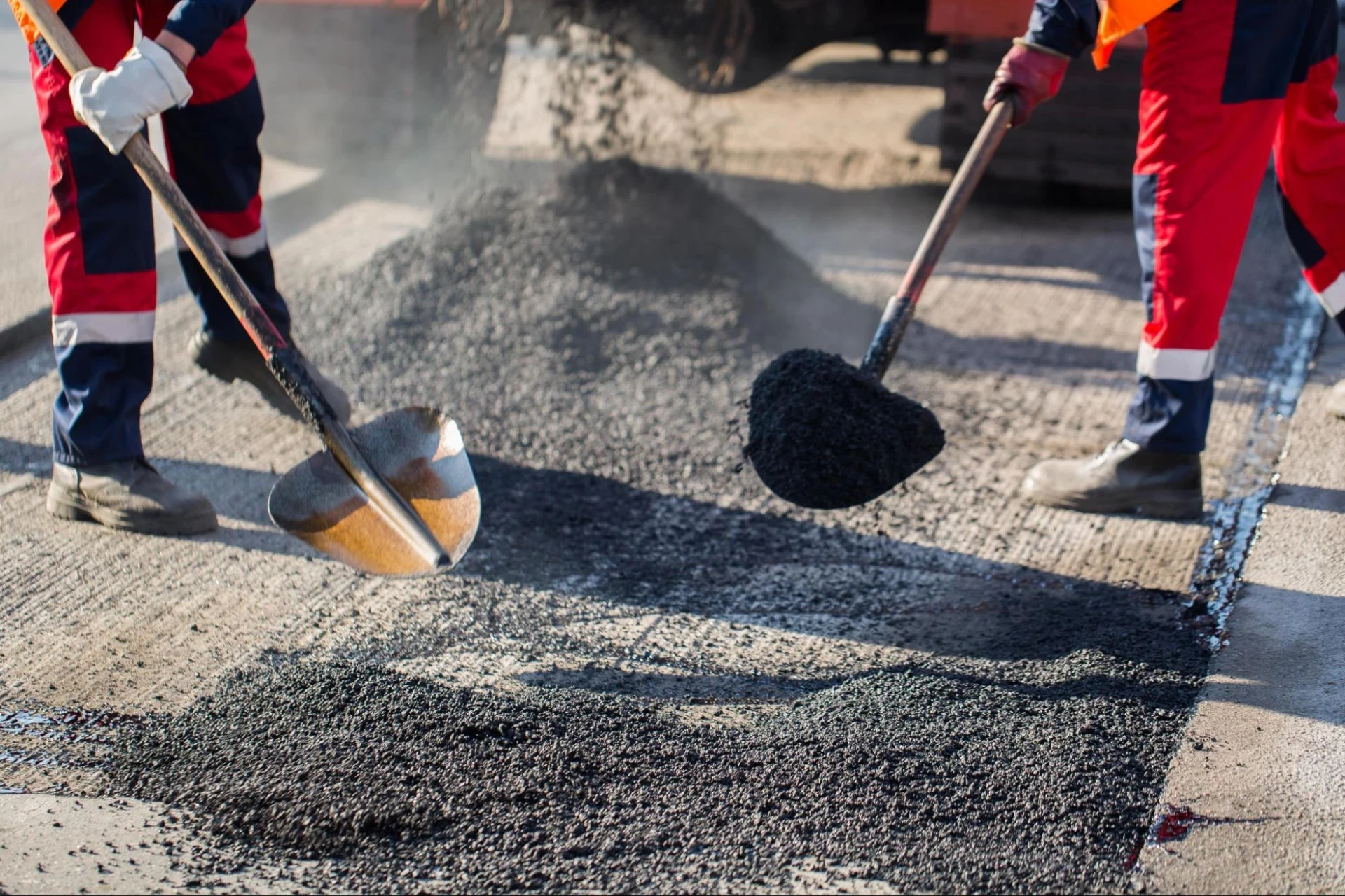 Workers in safety gear are laying asphalt on a road using shovels.