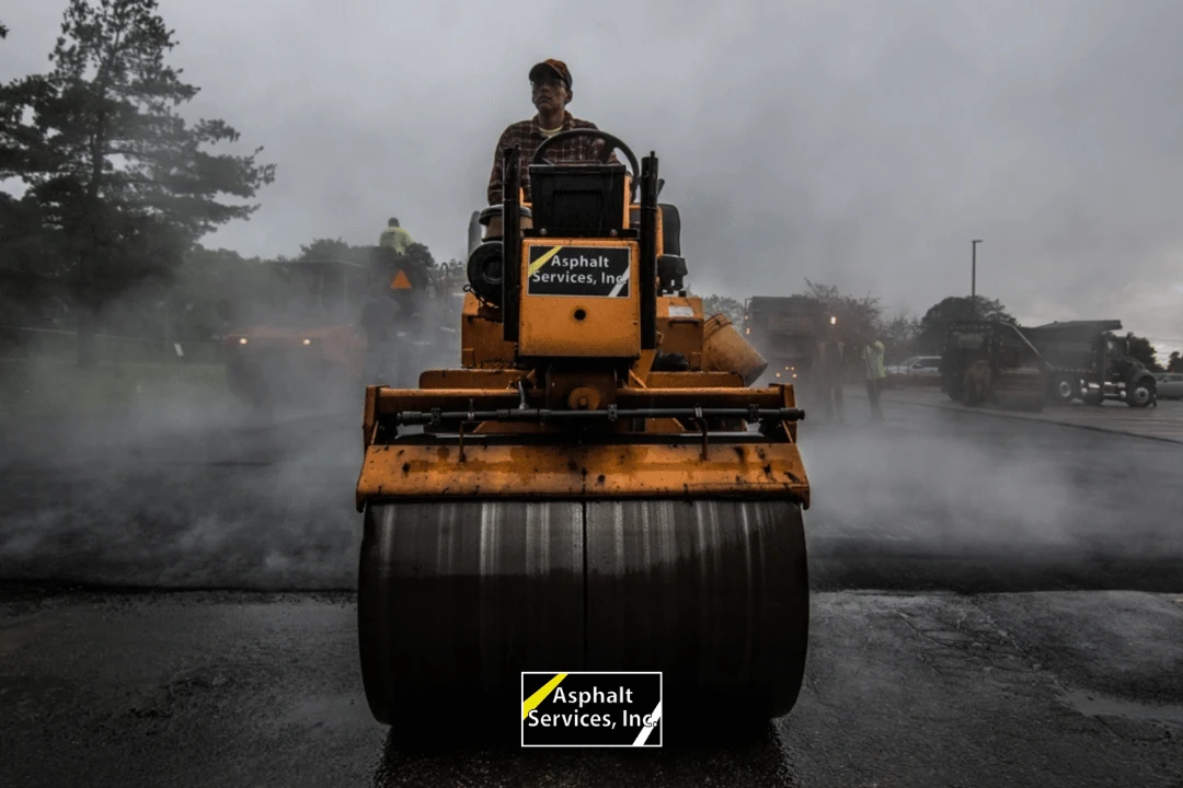 A worker operates a yellow asphalt roller on a steaming road, with "Asphalt Services, Inc." displayed on the machine; other workers and machinery are seen in the misty background.