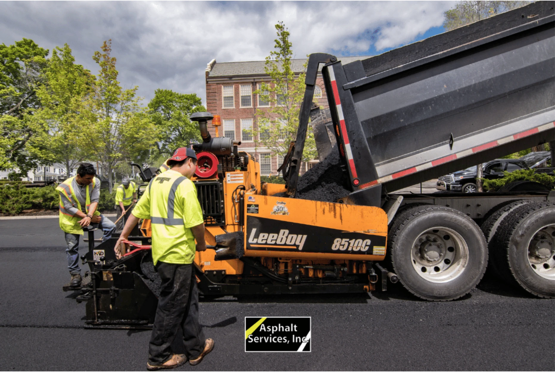 Workers of top paving contractor Asphalt Services Inc. operate paving equipment next to a dump truck pouring asphalt onto a road in front of a brick building, with trees and greenery in the background.