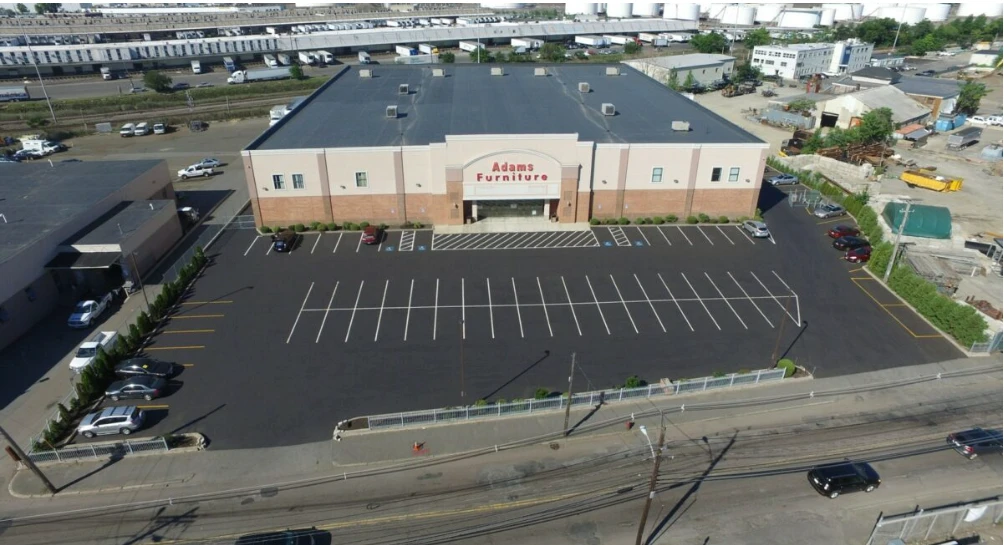 Aerial view of Adams Furniture store with an empty, freshly paved asphalt parking lot; surrounding area includes industrial and commercial buildings.