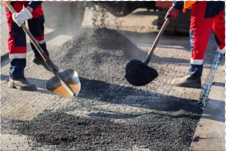 Workers in safety gear use shovels to spread fresh asphalt on a road surface during daylight, ensuring even coverage, a process often estimated beforehand with an Asphalt Driveway Cost Calculator.