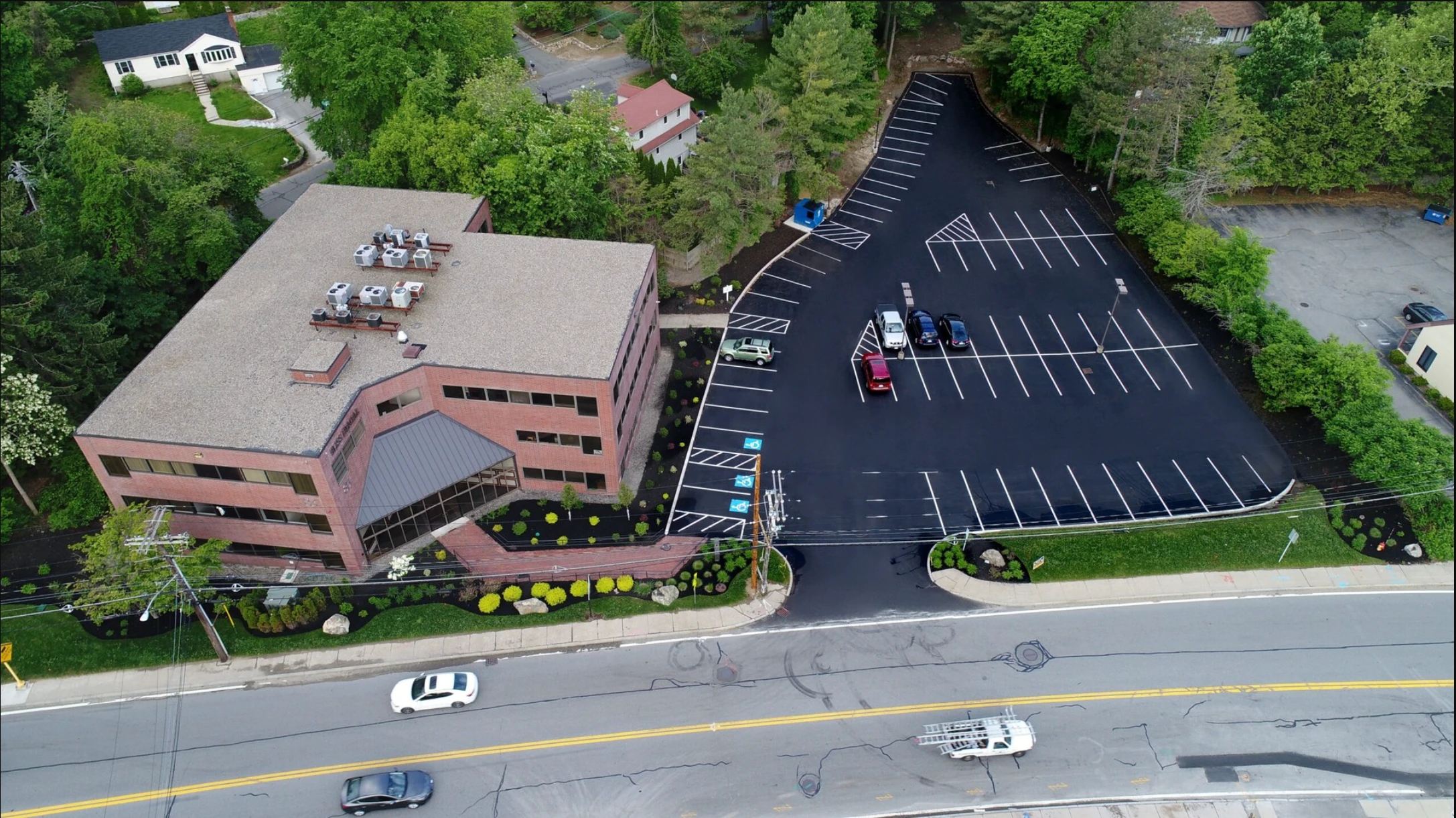 Aerial view of a brick office building with a parking lot, marked spaces, several parked cars, and surrounding greenery along a main road. The asphalt is freshly paved, and will not need parking lot restriping for some tim.
